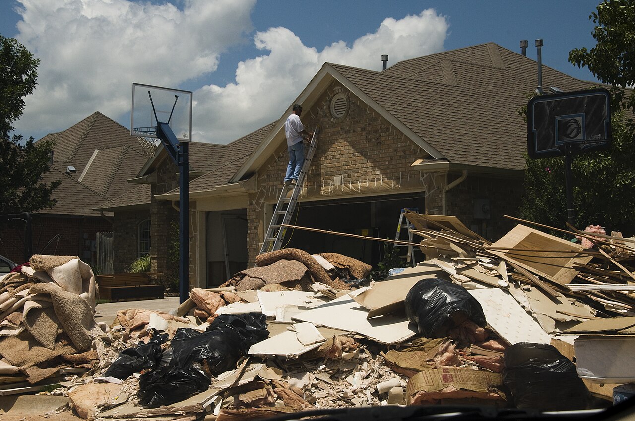 A homeowner on a ladder repairs a flood-damaged brick house while removed carpet, drywall, and debris sit piled at the curb.