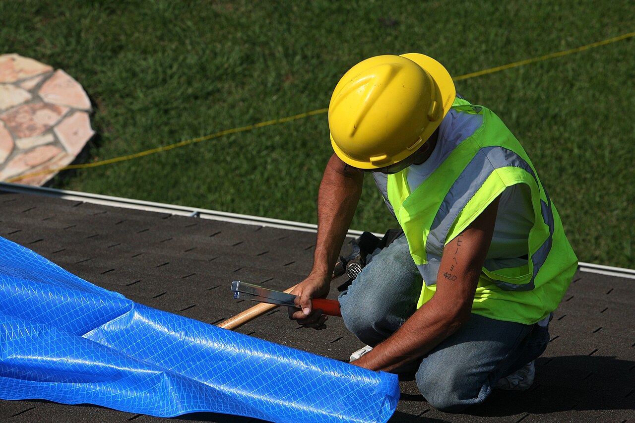 A worker fastens a blue emergency tarp over the shingles of a storm-damaged roof.